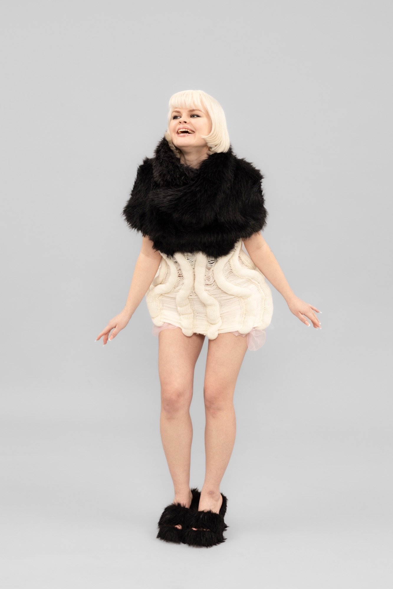 Lookbook: Model lachend vor grauem Grund, in Strickkleid mit schwarzer Fellstola und Plateau-Fellsandalen